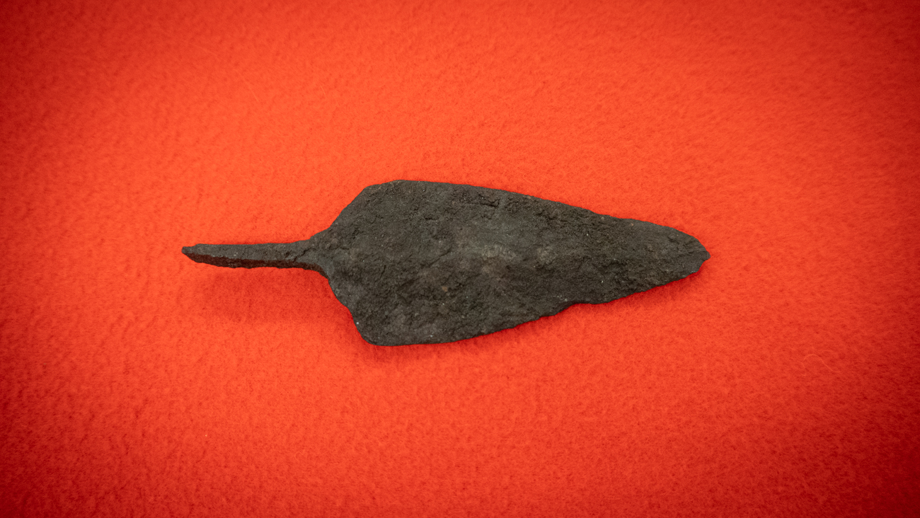 Rautanuolenkärki Juuan Nevalasta rautakauden alkupuolelta, 200-600-luvulta. Nuolenkärki on varrellinen ja väriltään tumma. Taustalla on punainen kangas. Kuva: Henna Palovaara.
