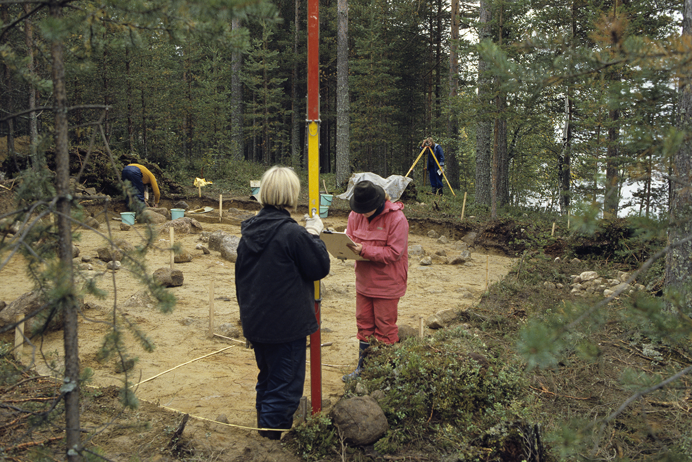Vaaitusta Ilomantsin Karpankankaalla vuonna 1987. Kaksi henkilöä mittaa maanpinnan korkeuseroja vaaittamalla. Taustalla arkeologit työskentelevät kaivauksella. Kuva: Museovirasto.