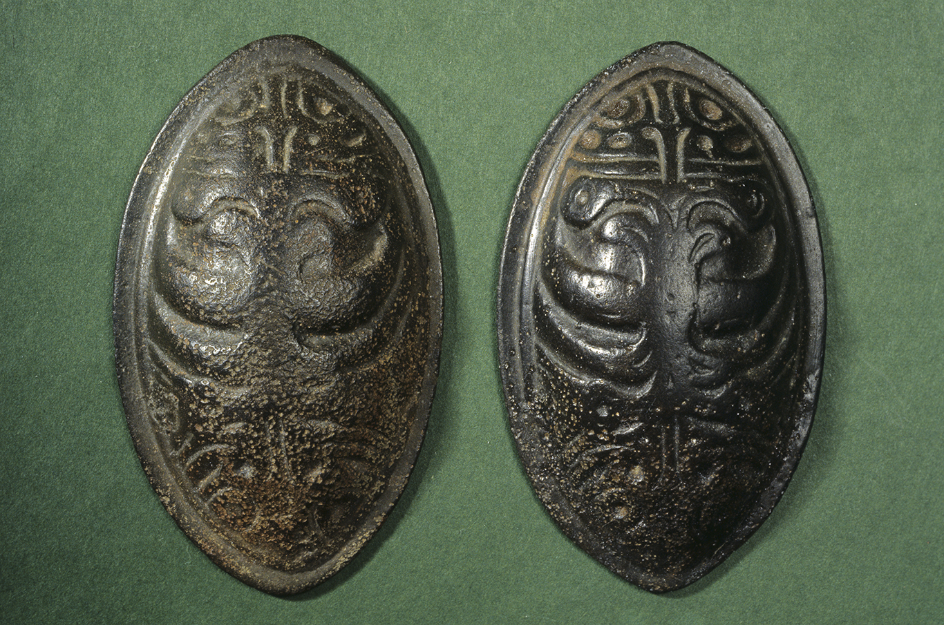 Kaksi soikeaa kupurasolkea Ilomantsista. Kupurasoljet ovat metallisia, koristeellisia soikeanmuotoisia solkia vihreällä taustalla. Kuva: Museovirasto.