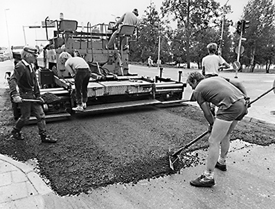 Mustavalkoinen kuva, jossa näkyy asfalttityöntekijöitä työn touhussa.